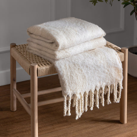 Meta Jacquard Polyester Throw in natural cream, folded with matching tasseled throw draped over a woven bench in a bedroom setting.