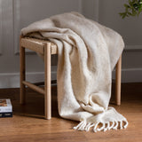 Meta Jacquard Polyester Throw in natural draped over a wooden bench with woven seat, showing soft brushed texture and twisted tassel fringe in a living room setting.
