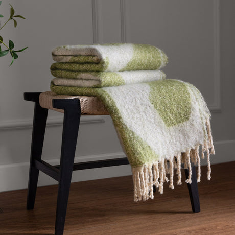 Meta Jacquard Polyester Throw in Olive, folded and draped with fringed edges on a black stool, showing its abstract green and cream woven pattern in a living room setting.