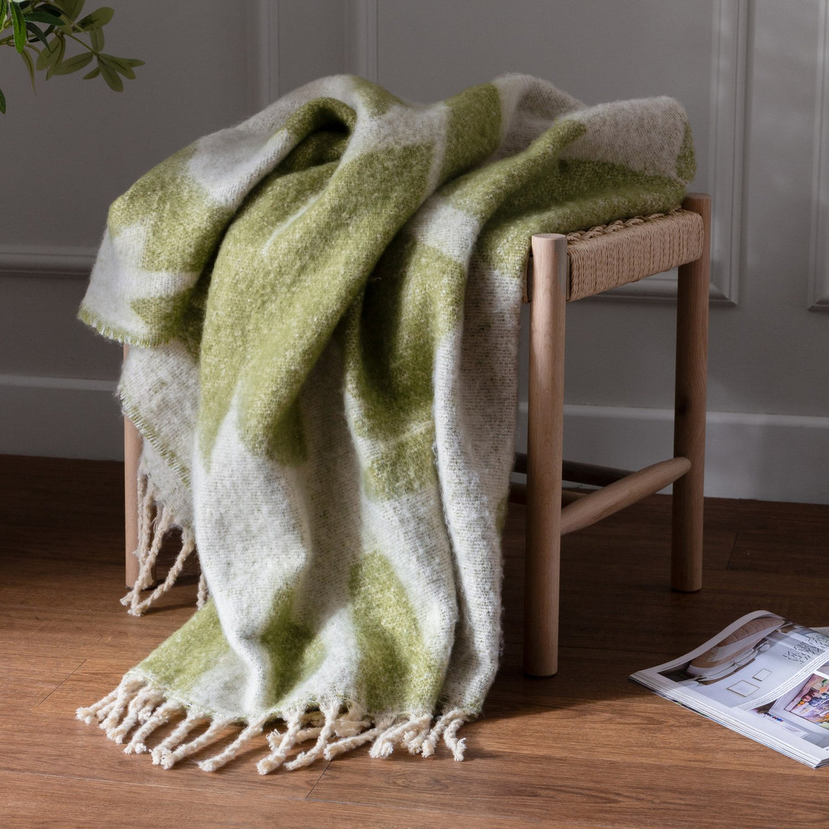 Meta Jacquard Polyester Throw in olive green and cream, draped over a wooden stool with a woven seat, showing a jacquard pattern and fringed edge.
