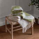 Meta Jacquard Polyester Throw in olive green and cream, folded with a fringed edge cream throw on a woven wooden stool in a living room setting.