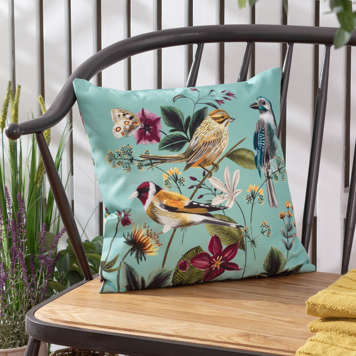 Square aqua polyester outdoor cushion with painted goldfinch, reed bunting and blue jay among floral botanicals, shown on a metal-framed bench.