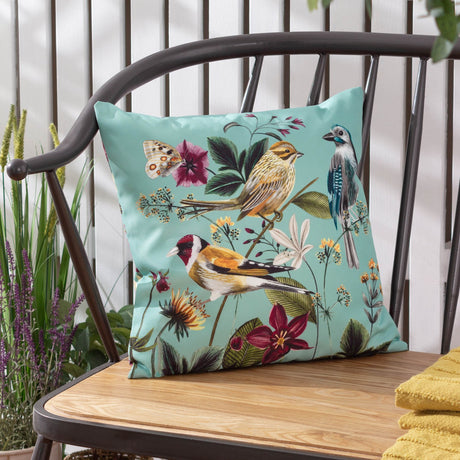 Square aqua polyester outdoor cushion with painted goldfinch, reed bunting and blue jay among floral botanicals, shown on a metal-framed bench.