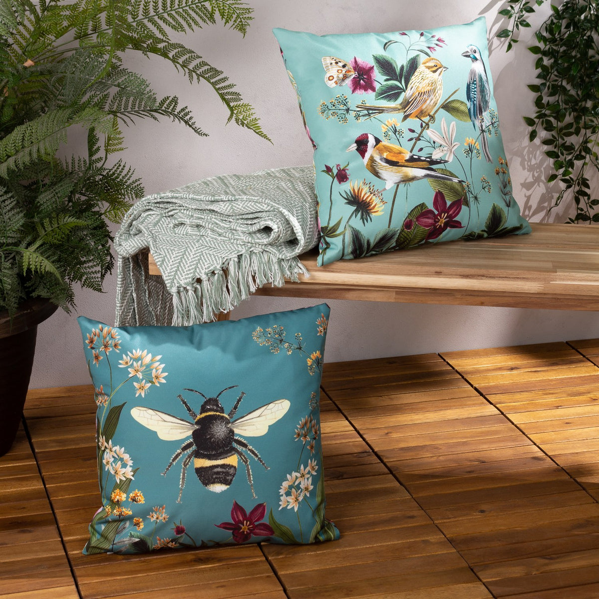 Aqua polyester outdoor cushion with bird and floral print shown on a wooden bench beside a matching bee-print cushion, throw, and potted plants.