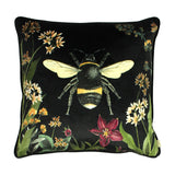 Square black velvet cushion featuring a large printed bumblebee, bordered by botanical stems, cream and amber blooms, and a red flower, shown on a plain white background.