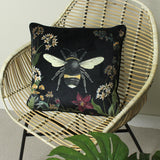 Midnight Garden Animal Velvet Cushion in black, square with a bumblebee design and floral border, shown on a rattan chair.