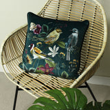 Midnight Garden Animal Velvet Cushion in green, printed with garden birds and florals with a piped edge, shown on a rattan chair.