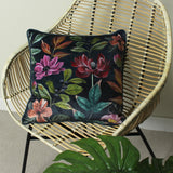 Midnight Garden Floral Velvet Cushion in grey with pink, crimson and coral blooms on green foliage, shown on a rattan chair beside a monstera leaf.