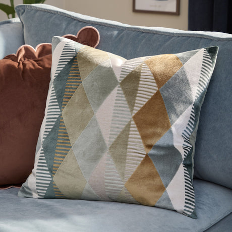 Metro Harlequin Jacquard Velvet Cushion in green, showing a diamond patchwork of cut-velvet and striped panels, styled on a blue sofa with a rust-pink cushion nearby.