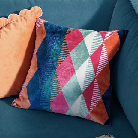 Metro Harlequin Jacquard Velvet Cushion in multicoloured, square with diamond pattern in pink, orange, teal, blue, cream and green, shown on a teal sofa beside an orange scalloped cushion.