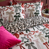 Mila Bows reversible duvet cover set on a bed, one side printed with red, green and cream bow motifs, reversing to a black and tan leopard print, styled with matching pillowcases and a pink cushion.
