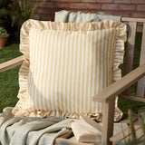 Millie Ruffle Striped Polyester Cushion in yellow and white, square with gathered ruffle trim, shown on an outdoor wooden bench beside a throw and book.