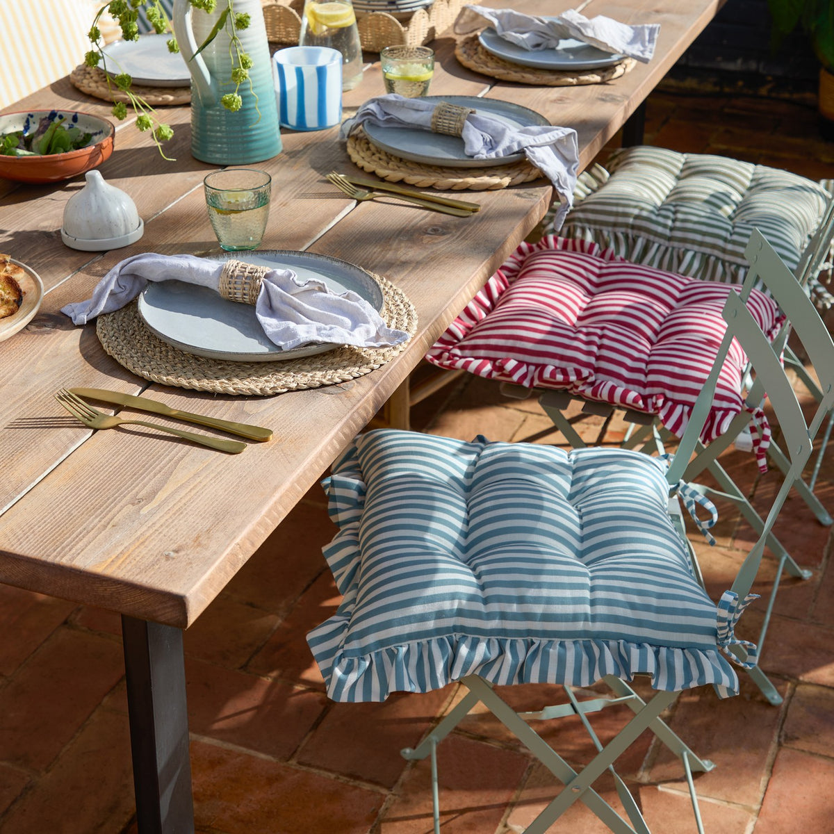 Millie Ruffle Striped Polyester Seat Pad in blue and white on an outdoor folding chair, with matching red and green striped pads on chairs beside a wooden dining table set for a meal.