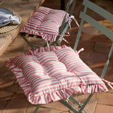 Millie Ruffle Striped Polyester Seat Pads in pink and white, tufted with ruffled edges, shown tied onto metal bistro chairs at an outdoor dining table.