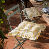 Millie Ruffle Striped Polyester Seat Pads in yellow and white, tied onto green metal bistro chairs at an outdoor dining table with a ruffled edge trim.