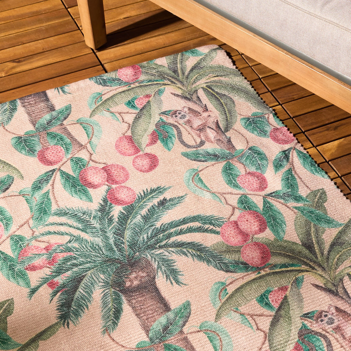 Monkey Palm Jungle polyester rug in papyrus, printed with palm trees, pink fruit and monkeys, shown on a wooden deck beside outdoor furniture.