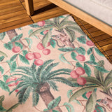 Monkey Palm Jungle polyester rug in papyrus, printed with palm trees, pink fruit and monkeys, shown on a wooden deck beside outdoor furniture.