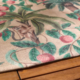Close-up of the Monkey Palm Jungle polyester rug in papyrus, showing a printed tropical scene with palm leaves, pink fruit and a monkey, edge resting on a wooden floor.