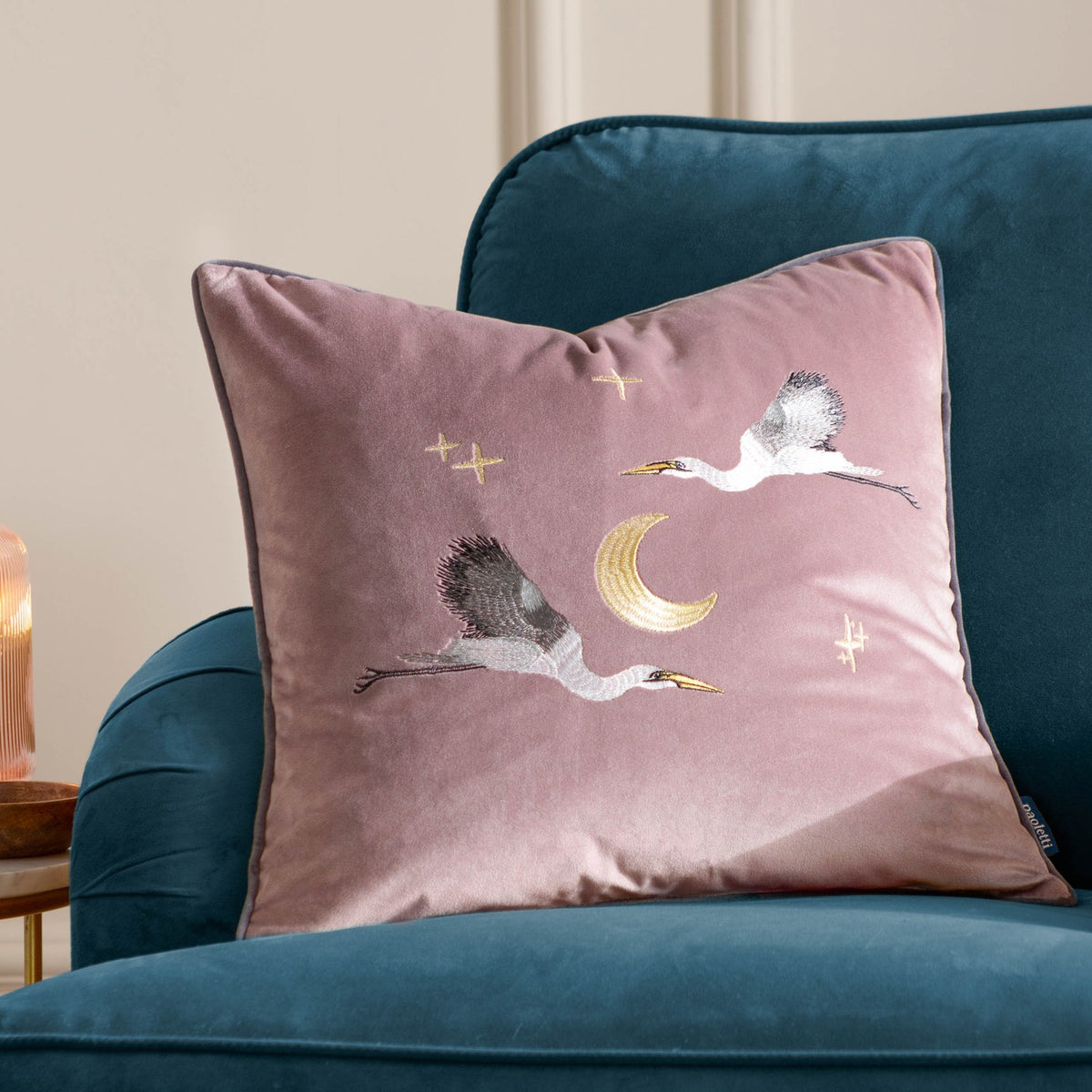 Dusty pink velvet square cushion embroidered with two flying storks, a gold crescent moon and stars, shown on a teal armchair.