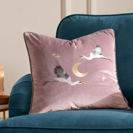 Dusty pink velvet square cushion embroidered with two flying storks, a gold crescent moon and stars, shown on a teal armchair.