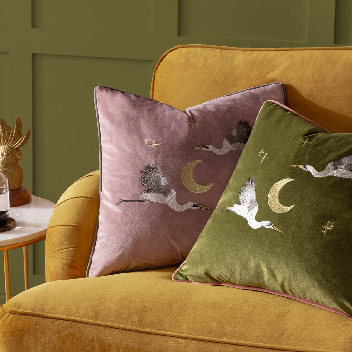 Moonlit Flight dusty pink velvet cushion with embroidered storks, crescent moon and stars, shown on a mustard sofa beside a matching olive green cushion.
