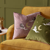 Moonlit Flight dusty pink velvet cushion with embroidered storks, crescent moon and stars, shown on a mustard sofa beside a matching olive green cushion.