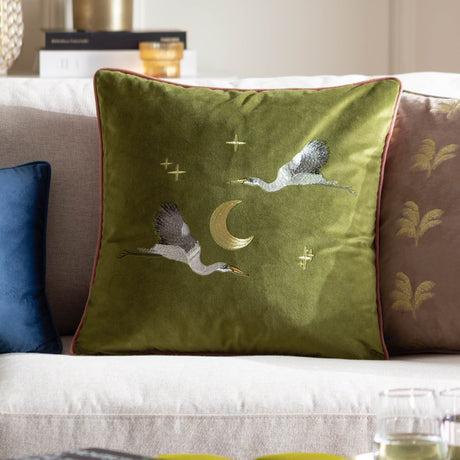 Moonlit Flight olive green velvet cushion with embroidered cranes, crescent moon and stars, pink piped edge, shown on a sofa beside patterned and blue cushions.