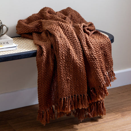 Morni Plain Polycotton Throw in Hazel, a chunky basketweave brown throw with tasselled fringe, draped over a bench beside stacked books in a living room setting.