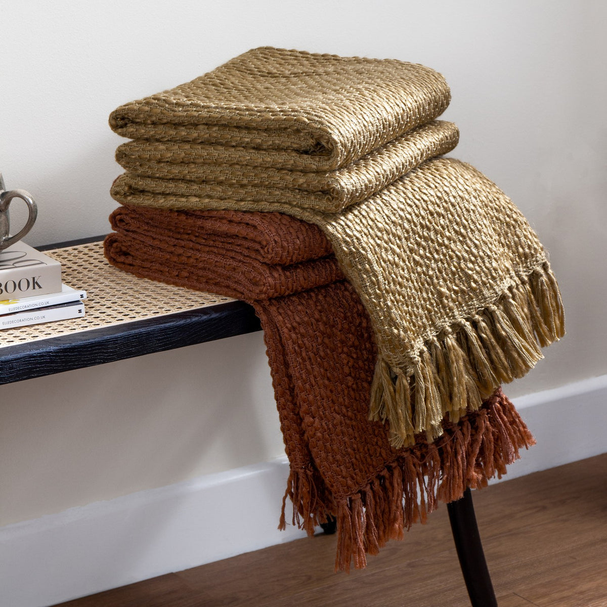Morni Plain Polycotton Throws in hazel gold and rust, folded with tasselled fringe edges, stacked on a bench in a living room setting.