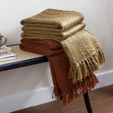 Morni Plain Polycotton Throws in hazel gold and rust, folded with tasselled fringe edges, stacked on a bench in a living room setting.