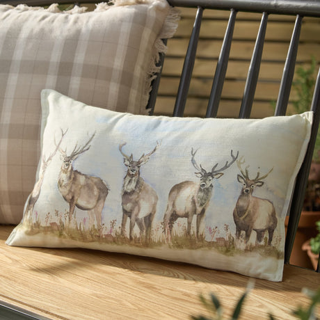 Moorland Stag Reversible Linen Cushion in Nut, rectangular cushion printed with watercolour stags on a meadow scene, shown on a bench beside a check-patterned pillow.