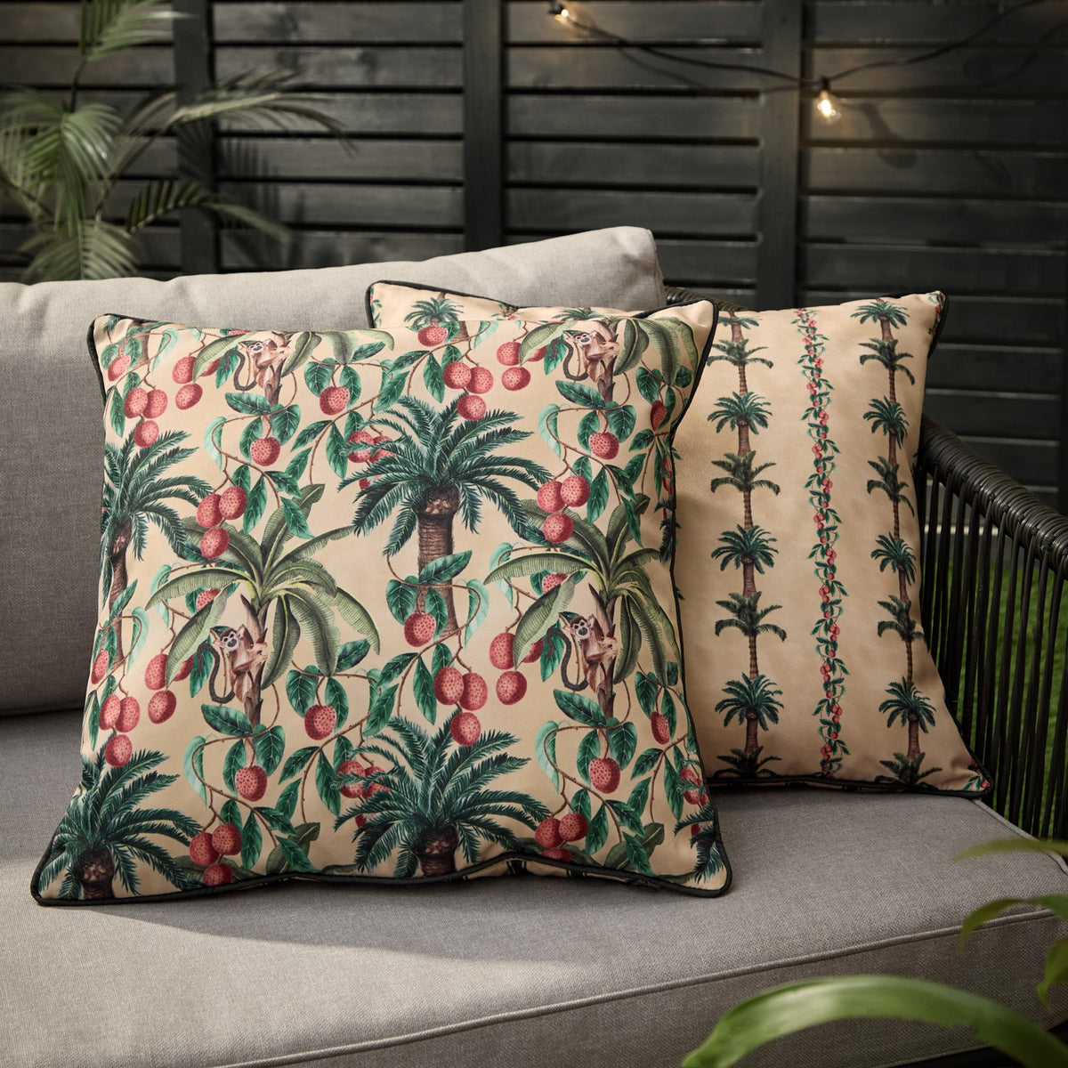 Reversible square outdoor cushions in beige with monkey and palm-tree print in green and coral-red, one showing a striped palm-tree side, on a grey garden sofa.