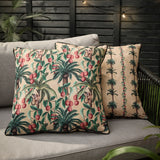 Reversible square outdoor cushions in beige with monkey and palm-tree print in green and coral-red, one showing a striped palm-tree side, on a grey garden sofa.