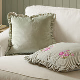 Martha Floral Linen Cushion in sage-and-cream ticking stripe with ruffled edge and pink floral embroidery, shown on a beige sofa beside a plain sage velvet cushion.