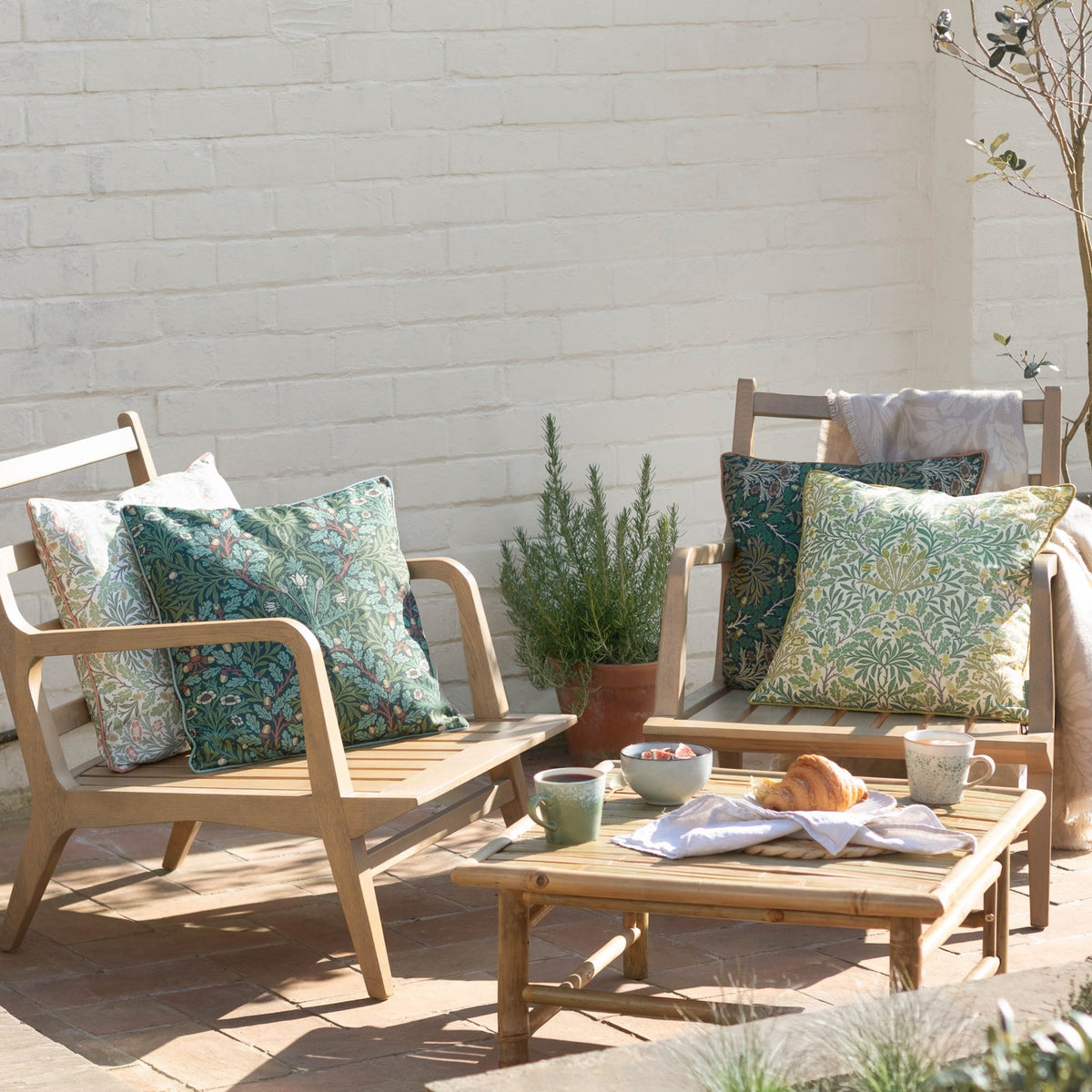 Multi Acorn Woodland polyester cushion in sage and stone green with cream piping, styled on wooden garden chairs alongside a matching cushion and outdoor seating area.