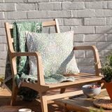 Multi Acorn Woodland polyester cushion in stone/sage on a wooden garden bench, with off-white ground, sage-green foliage, terracotta flowers and caramel piping, alongside a matching green throw.