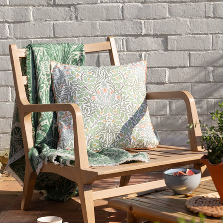 Multi Acorn Woodland polyester cushion in stone/sage on a wooden garden bench, with off-white ground, sage-green foliage, terracotta flowers and caramel piping, alongside a matching green throw.
