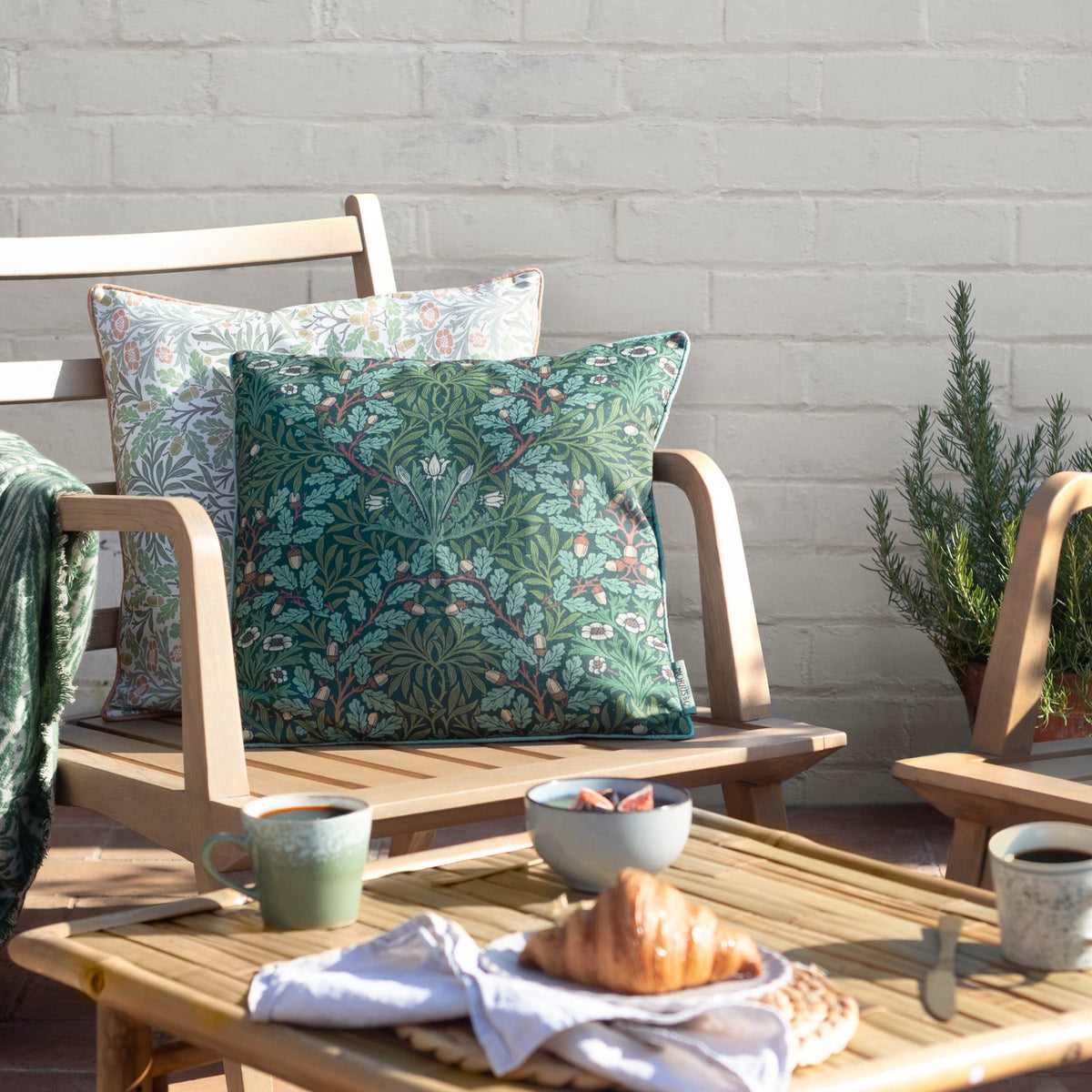 Multi Acorn Woodland polyester cushion in Thyme/Forest green, oak leaf and acorn print, styled on a wooden garden chair beside a matching floral cushion.