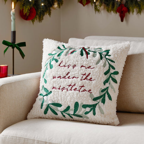 Cream boucle cushion on a sofa, embroidered with a green mistletoe wreath and "kiss me under the mistletoe" text, styled in a festive living room setting.