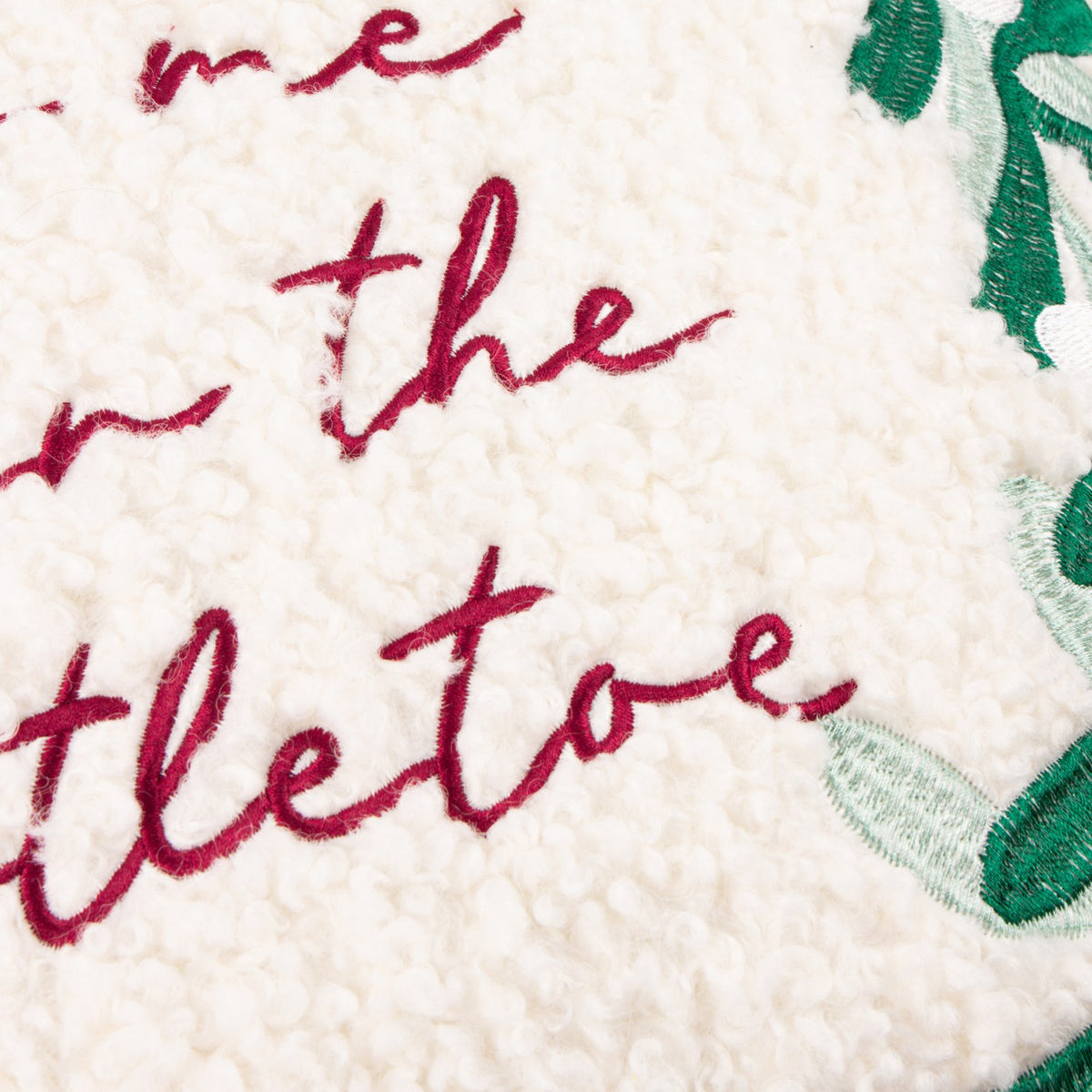 Close-up of the Mistletoe Wreath Embroidered Velvet Cushion, showing cream boucle fabric with "kiss me under the mistletoe" embroidered in red script beside green mistletoe leaves.