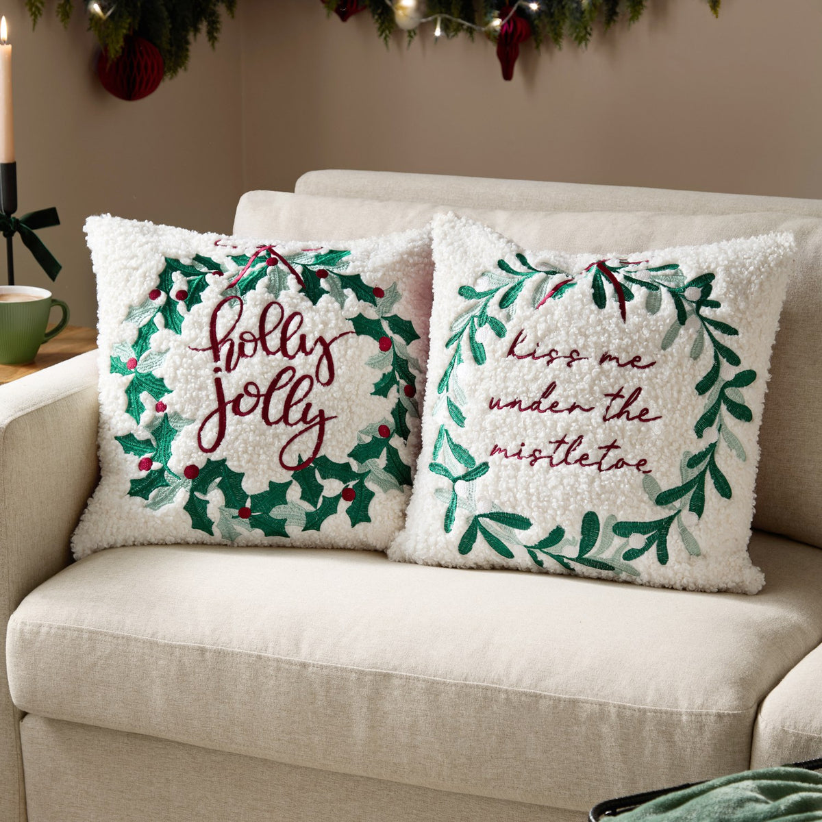 Boucle cushion with embroidered mistletoe wreath and "kiss me under the mistletoe" text, shown beside a matching "holly jolly" wreath cushion on a cream sofa.