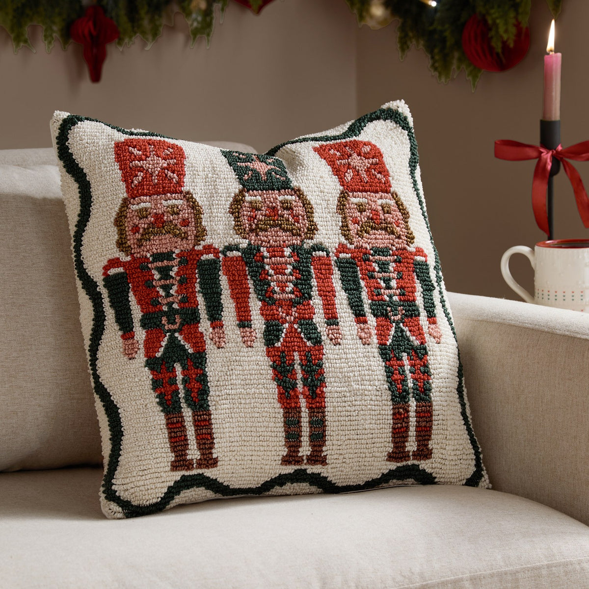 Square tufted cotton cushion featuring three nutcracker soldiers in red and green with a scalloped green border on a cream ground, shown on a sofa in a festive living room setting.