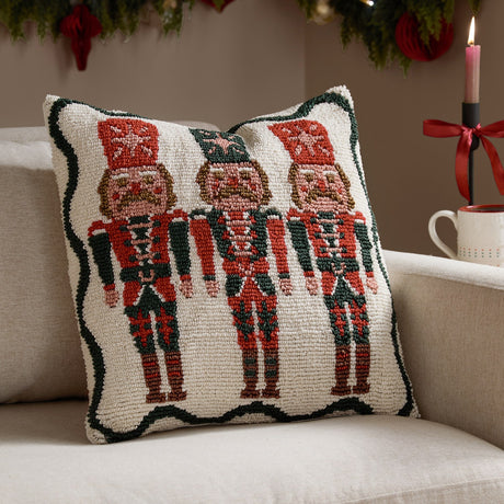 Square tufted cotton cushion featuring three nutcracker soldiers in red and green with a scalloped green border on a cream ground, shown on a sofa in a festive living room setting.