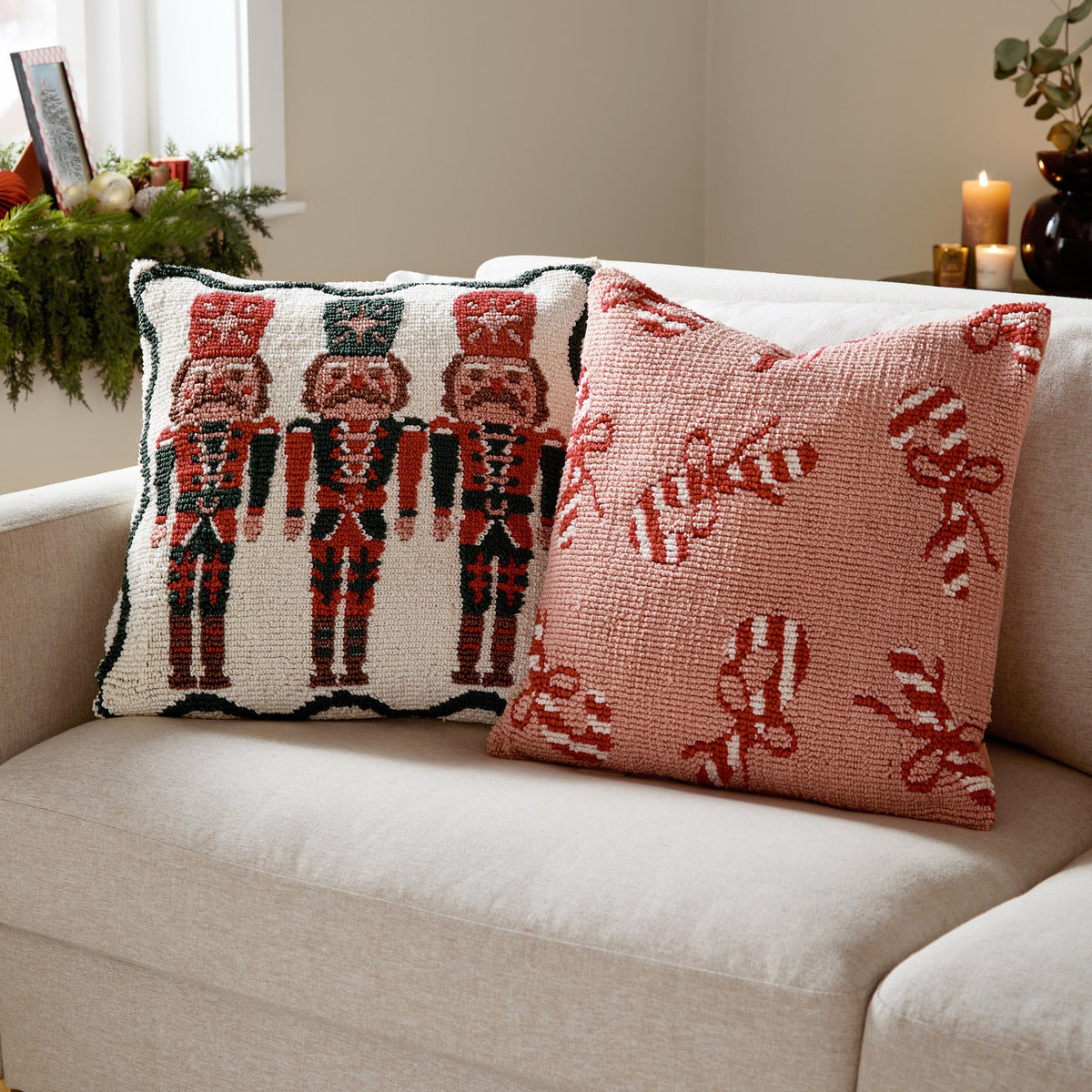 Nutcracker Trio Tufted Cotton Cushion in cream with three red and green nutcracker soldiers, shown on a sofa beside a pink candy cane and bow patterned cushion.