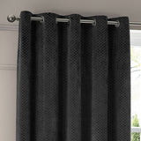 Neptune Velvet Fully Lined Eyelet Curtains in charcoal, showing textured honeycomb-patterned fabric with brushed metal eyelets on a silver pole beside a window.