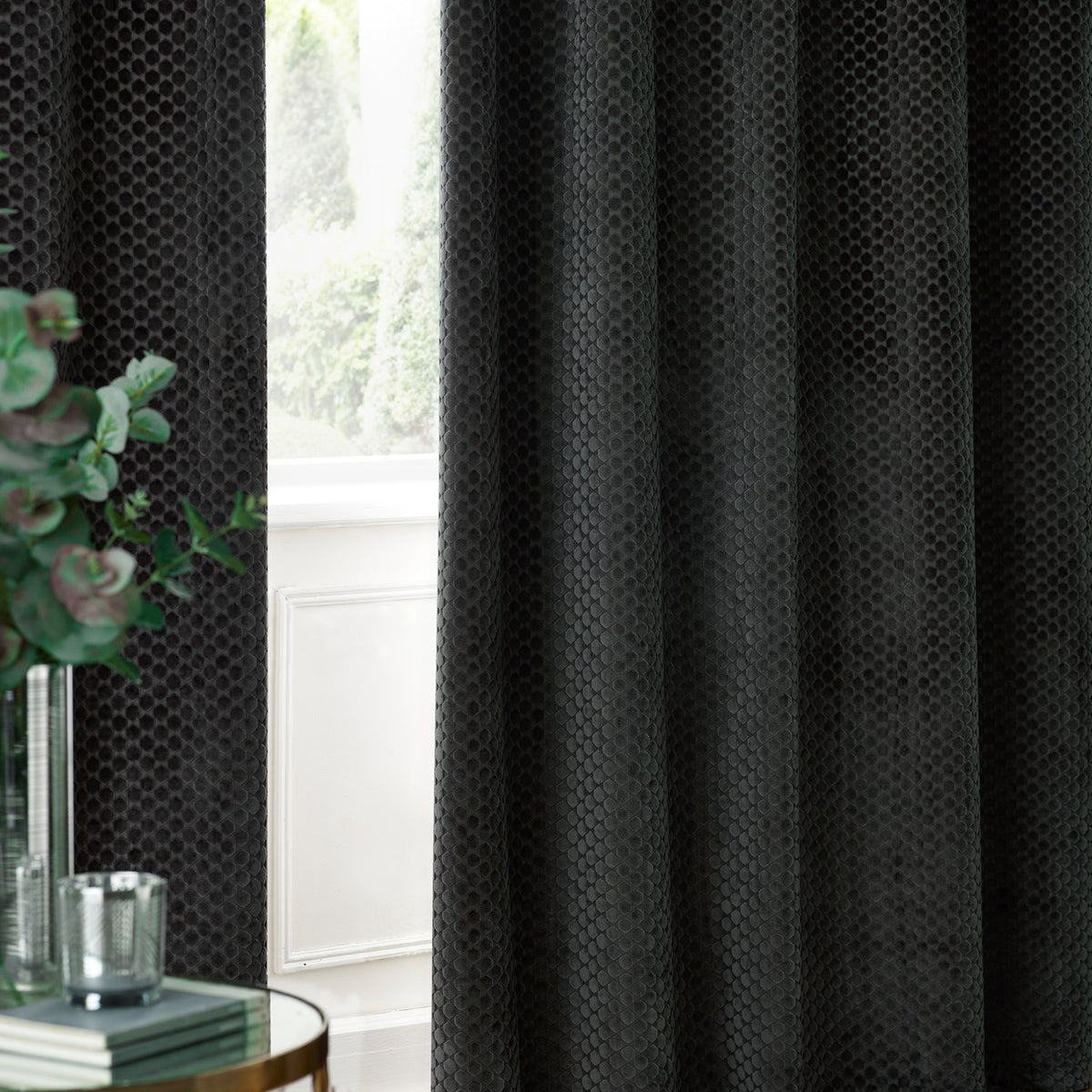 Neptune Velvet Fully Lined Eyelet Curtains in charcoal, showing raised honeycomb-textured fabric hung beside a window in a living room setting.