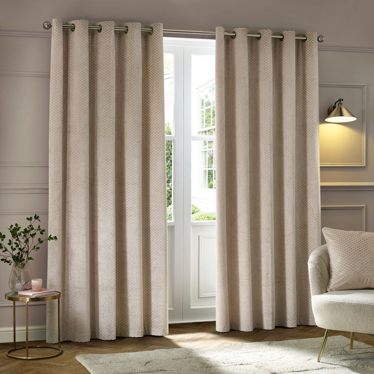 Neptune Velvet Fully Lined Eyelet Curtains in champagne, textured dot pattern fabric with metal eyelets, hung by a window in a living room setting.