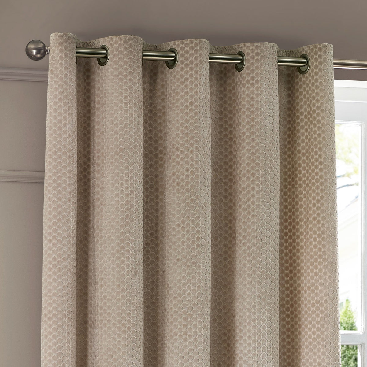 Neptune Velvet Fully Lined Eyelet Curtains in champagne, showing cut velvet fabric with a repeating geometric dot pattern and metal eyelet heading on a rod, hung in a living room.