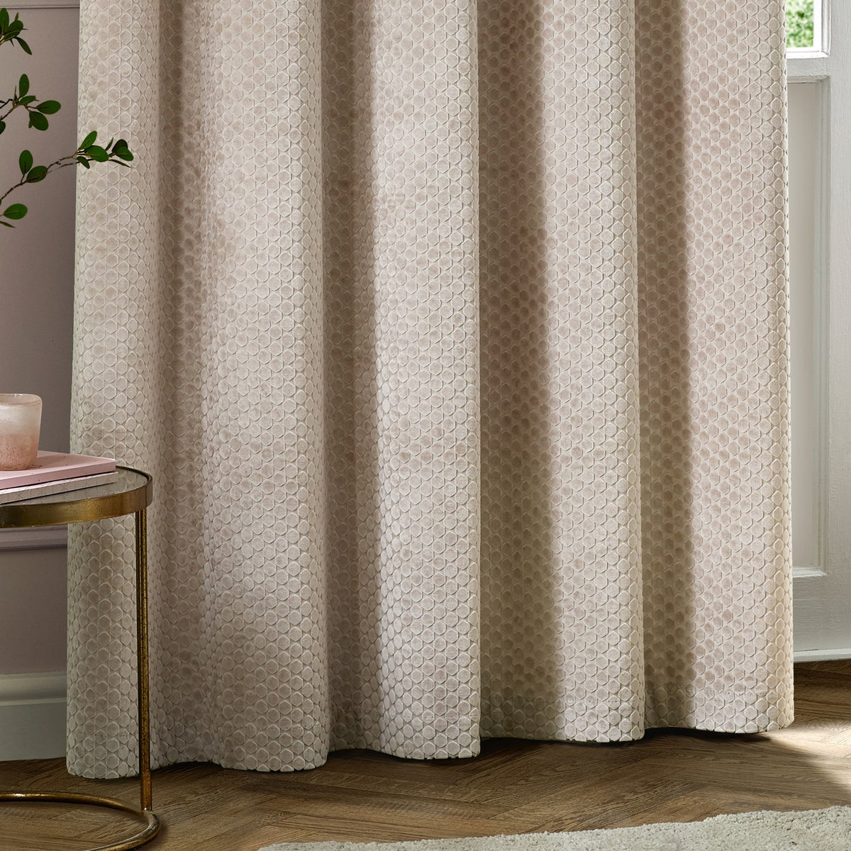 Neptune Velvet Fully Lined Eyelet Curtains in champagne, showing eyelet-header folds with a subtle dot-patterned cut velvet texture, styled in a living room with wood flooring.