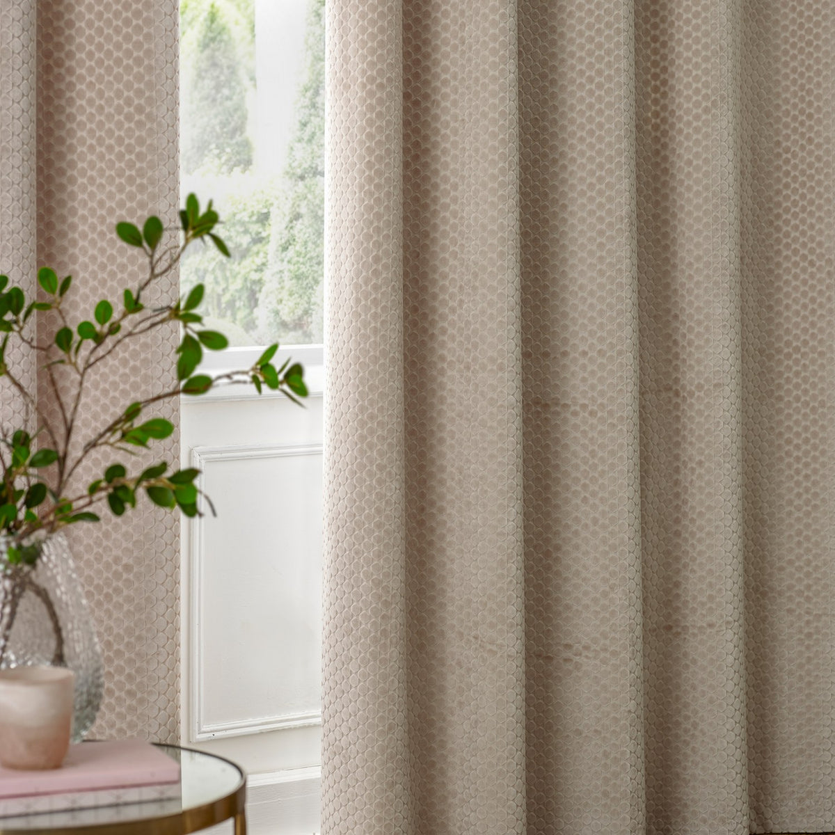 Neptune Velvet Fully Lined Eyelet Curtains in champagne, shown draped in soft folds with a subtle dot-textured pile, styled beside a bright window in a living room setting.